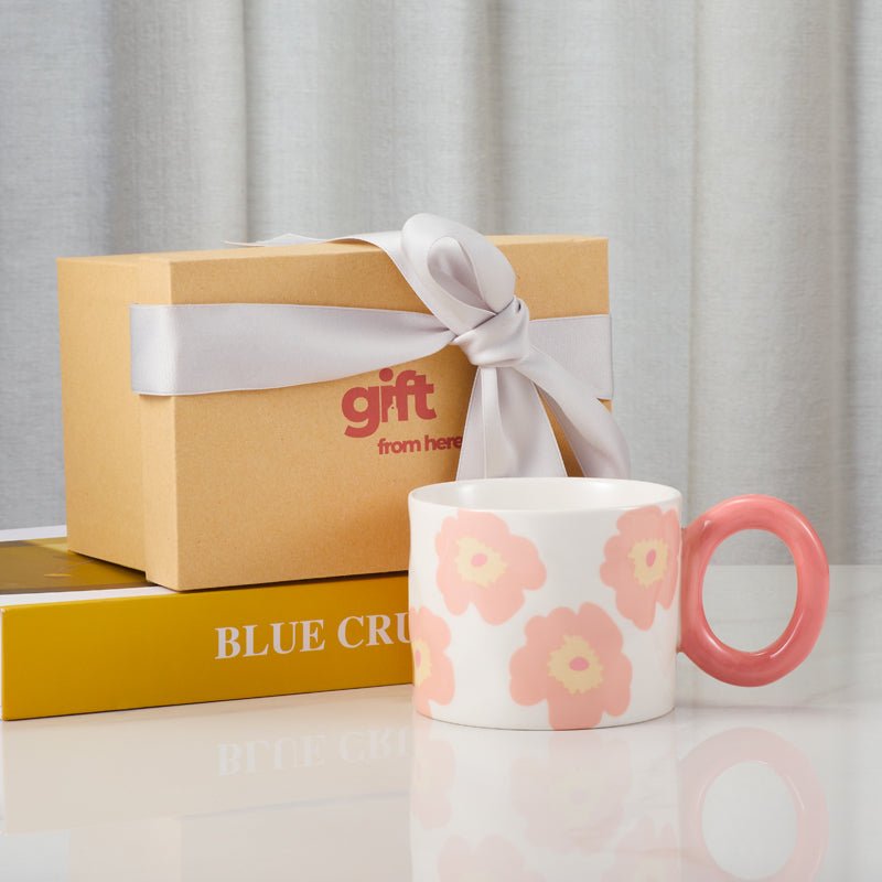 Floral-patterned ceramic mug with pink handle next to a gift box with a ribbon