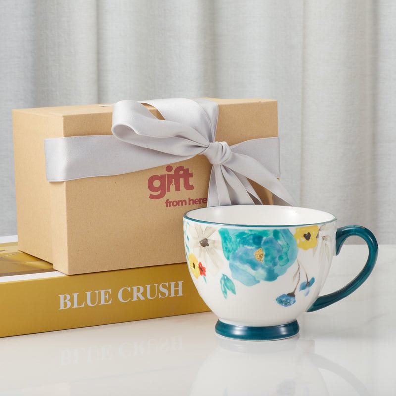 Floral-patterned ceramic mug with blue handle next to a gift box with a ribbon."